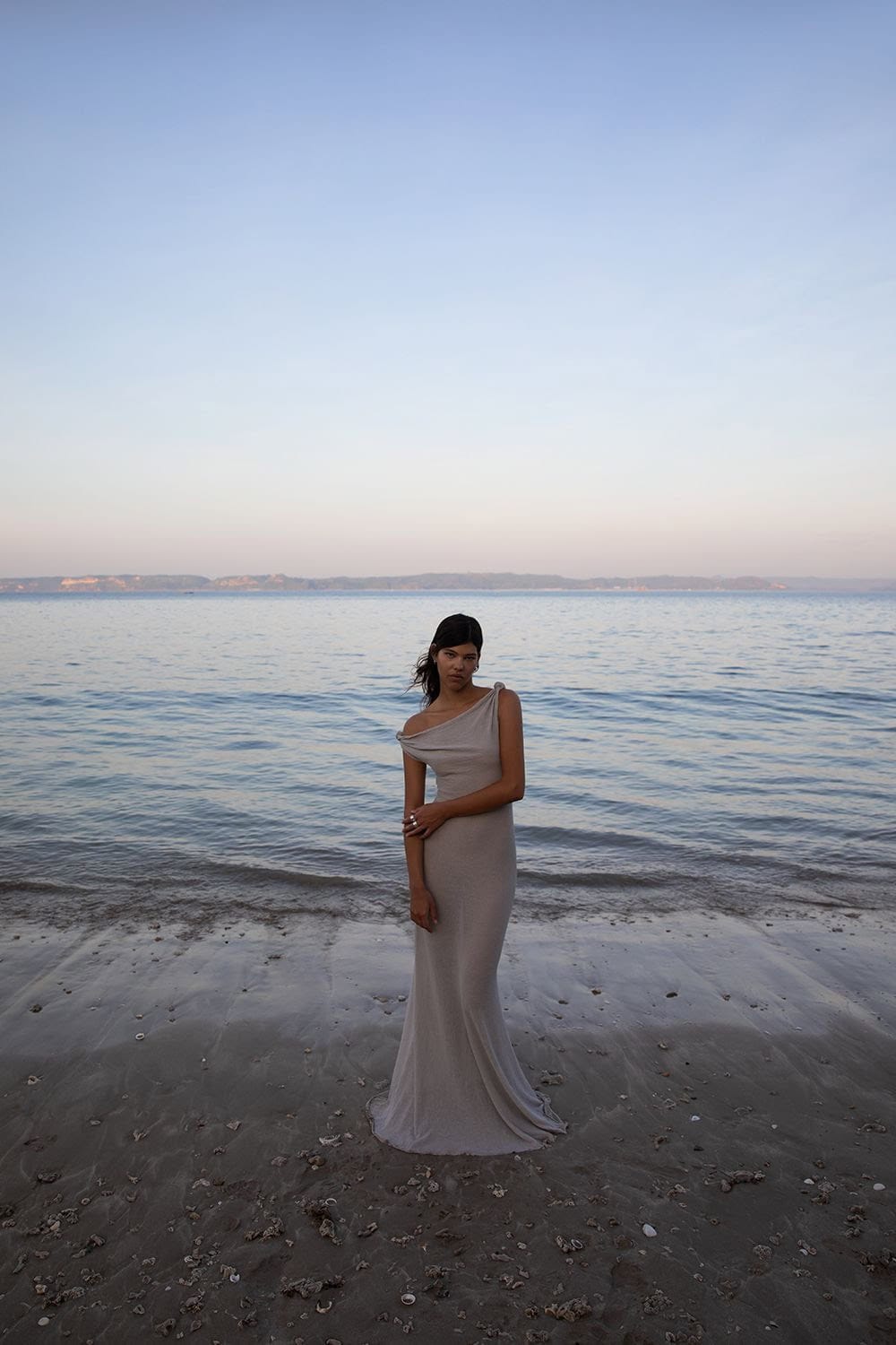 A woman in a gown stands on a sandy beach near the ocean at dusk, featuring soft lighting and a serene atmosphere. Dress is flowing.