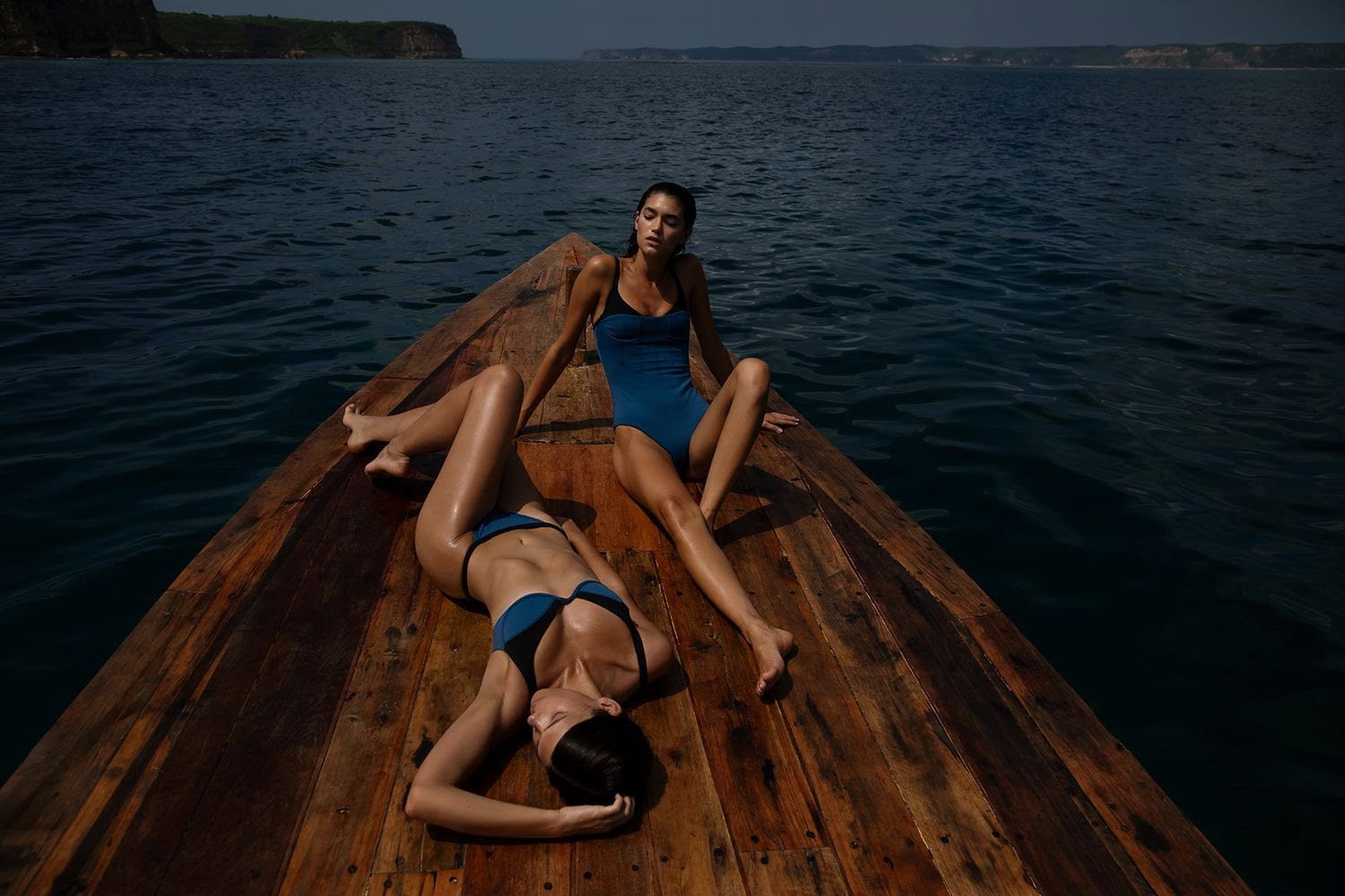 Two adult women in swimsuits lounge on a wooden boat floating on the ocean under daylight, with a distant landmass in the background.