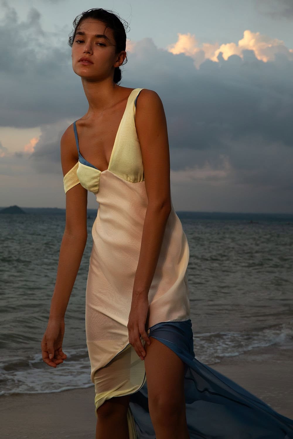 A woman in a draped gown stands on a beach with a sea and cloudy sky in the background, presenting a fashion or editorial aesthetic.