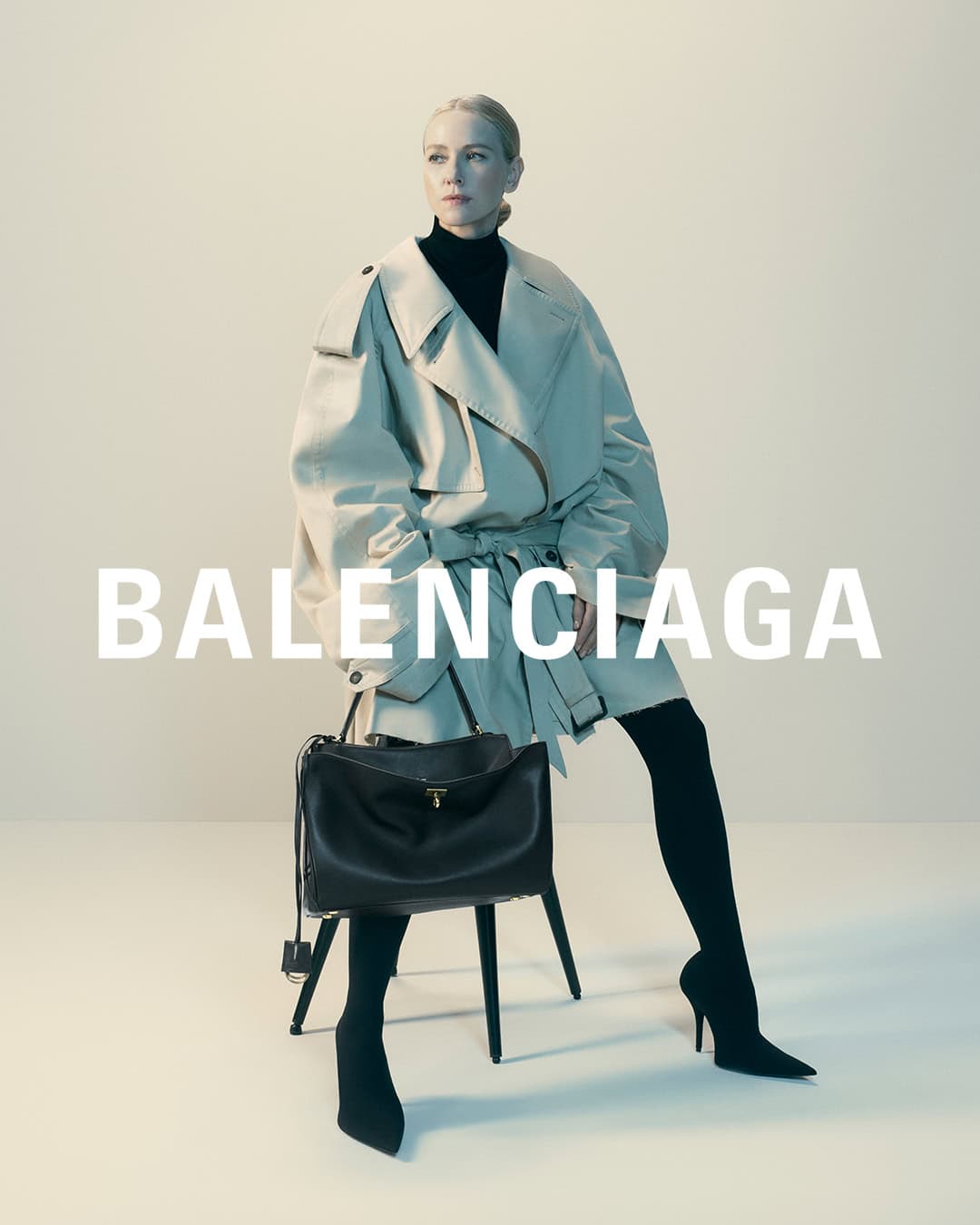 Blonde woman in a tan trench coat and black turtleneck poses with a black purse against a neutral backdrop for a Balenciaga fashion advertisement.