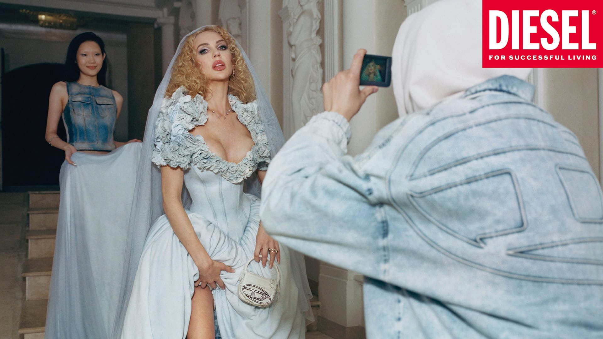 Fashion editorial image featuring a woman in a light-blue wedding gown with a Diesel handbag, being photographed indoors with another woman.