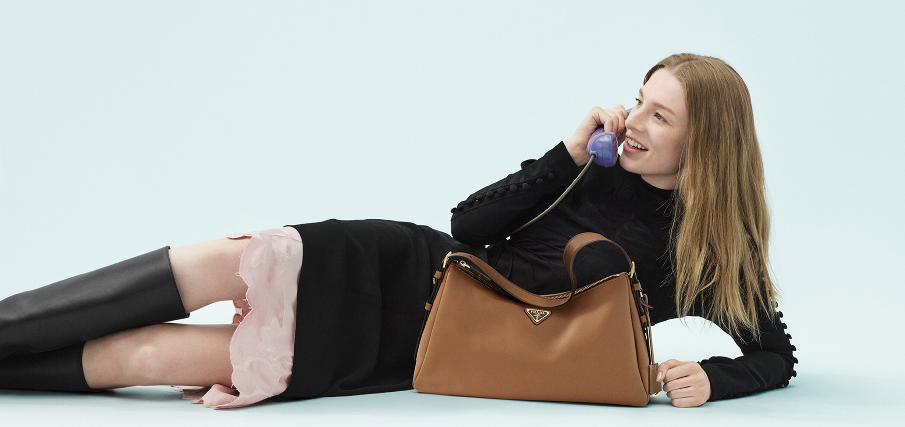 A woman in a black dress and boots reclines on a light blue surface, talking on a vintage-style blue rotary telephone, with a brown Prada bag.