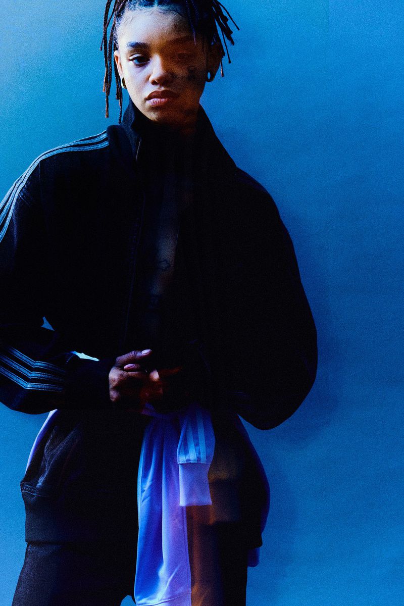 Portrait of a stylish woman with dreadlocks wearing a black Adidas jacket and a white and blue athletic shirt in a blue photo studio.