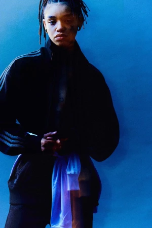 Portrait of a stylish woman with dreadlocks wearing a black Adidas jacket and a white and blue athletic shirt in a blue photo studio.