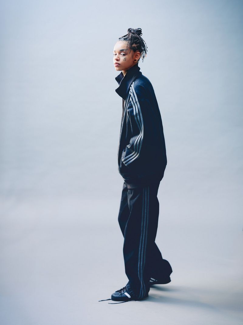 Full-length shot of a woman with braided hair posing in a navy blue and grey Adidas tracksuit in a light grey studio environment.