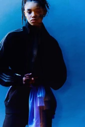 A young female model with dreadlocks wearing a black track jacket against a solid blue background for Exit Magazine.