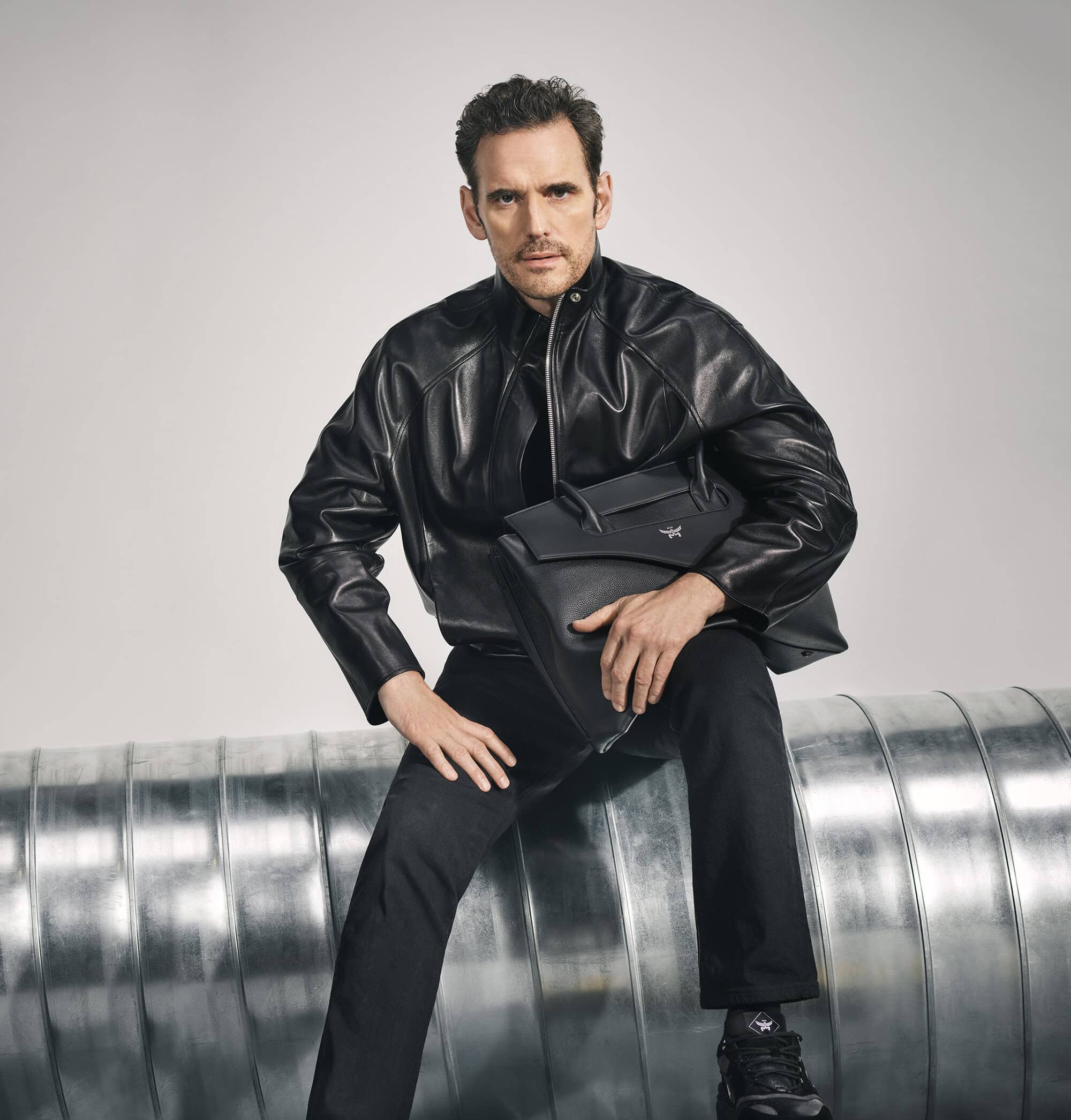 A man in a black leather jacket sits on a metal pipe holding a black bag in a studio with a neutral background.