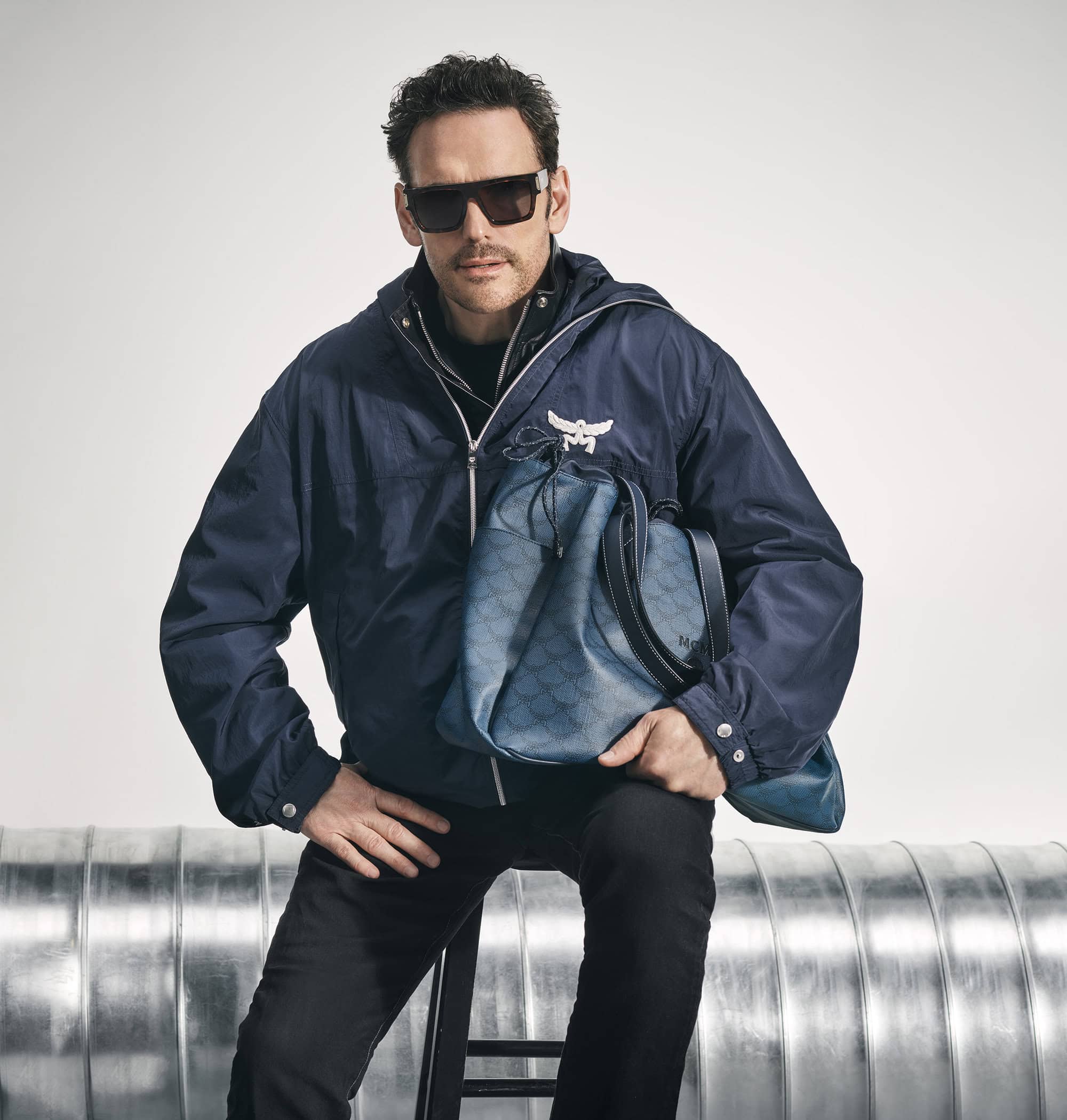 Adult male in sunglasses poses in a blue hooded jacket holding a blue patterned bag, seated against a metal pipe background.