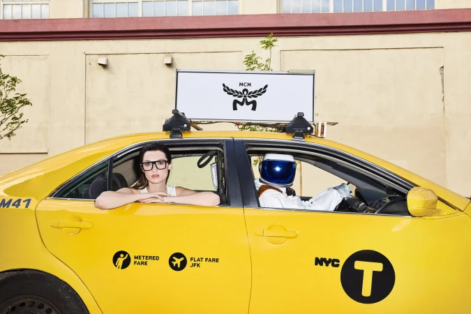 A woman leans out of a yellow taxi cab, with an astronaut driving. An MCM logo appears on a rooftop sign on the vehicle.