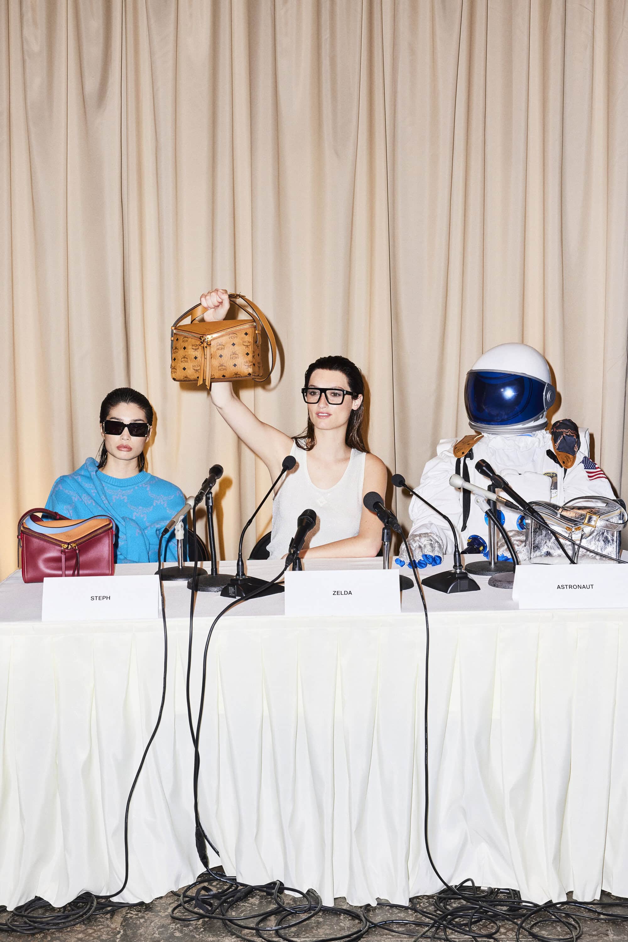 Two models and an astronaut sit at a press conference table showcasing designer purses in front of a tan curtain backdrop.