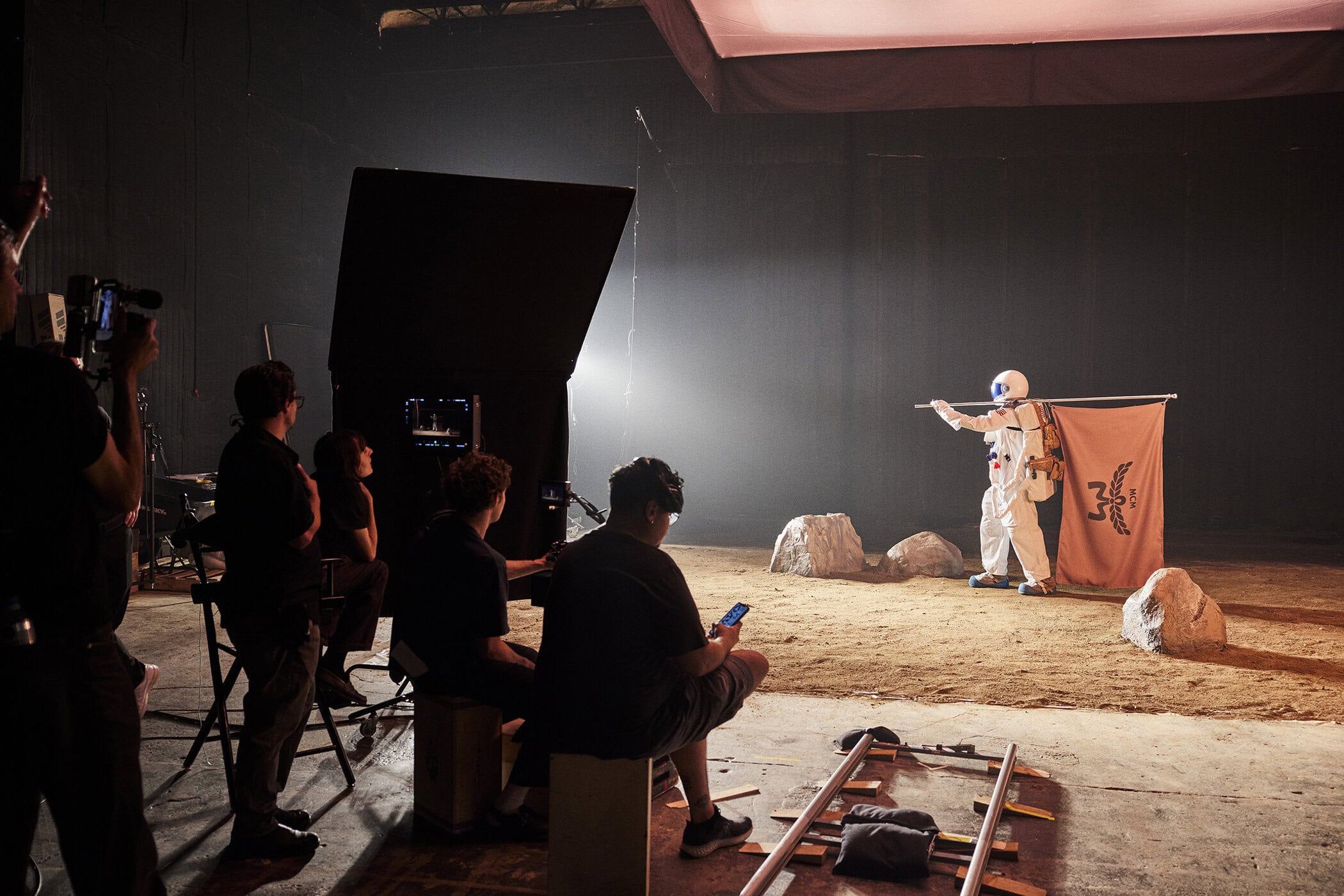 Astronaut on studio set with film crew. Man in space suit holds flag. Crew members watching on chairs and filming scene.