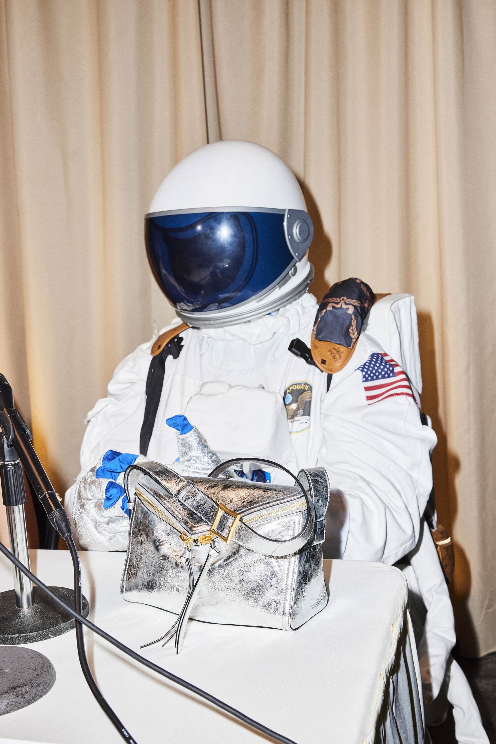 Person in a white spacesuit with a silver Loewe purse sitting at a table with a microphone, American flag patch on the shoulder.