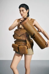 Model wearing multiple brown logo print bags and holding a cylindrical bag against a white seamless background