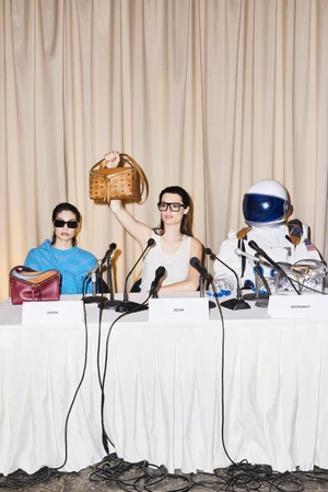 Three figures seated behind a draped press conference table holding brown leather handbags against a curtained backdrop.