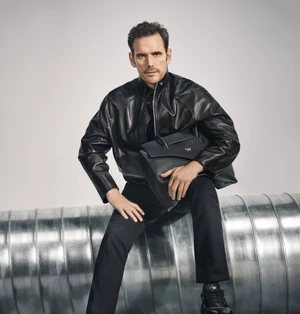 Matt Dillon wearing a black leather jacket and holding a black bag while seated on a metallic cylindrical structure