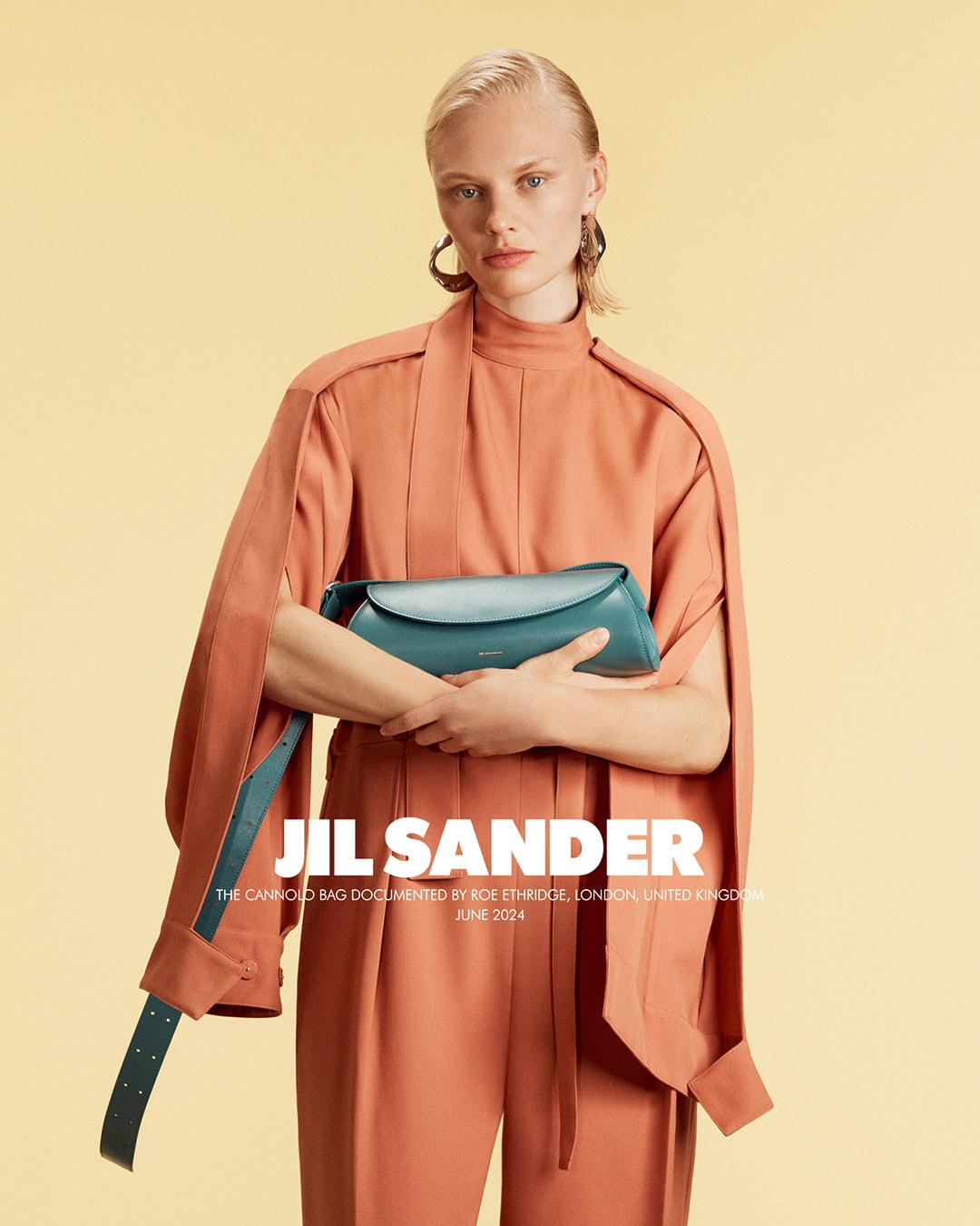 Fashion shot of a female model holding a blue handbag, wearing a peach-colored suit in front of a yellow backdrop in a studio.