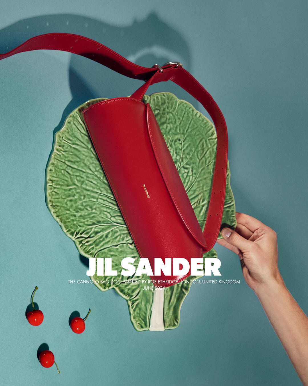 Red Jil Sander crossbody bag with strap displayed on a ceramic cabbage leaf with cherries against a light blue backdrop.