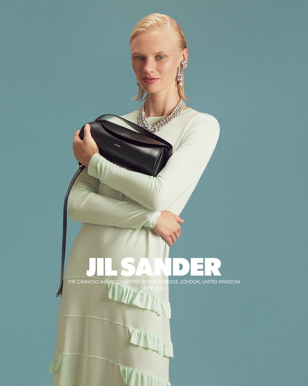 Fashion editorial shot of a blonde model posing with a black handbag, wearing a long light-green dress and jewelry against a teal background.