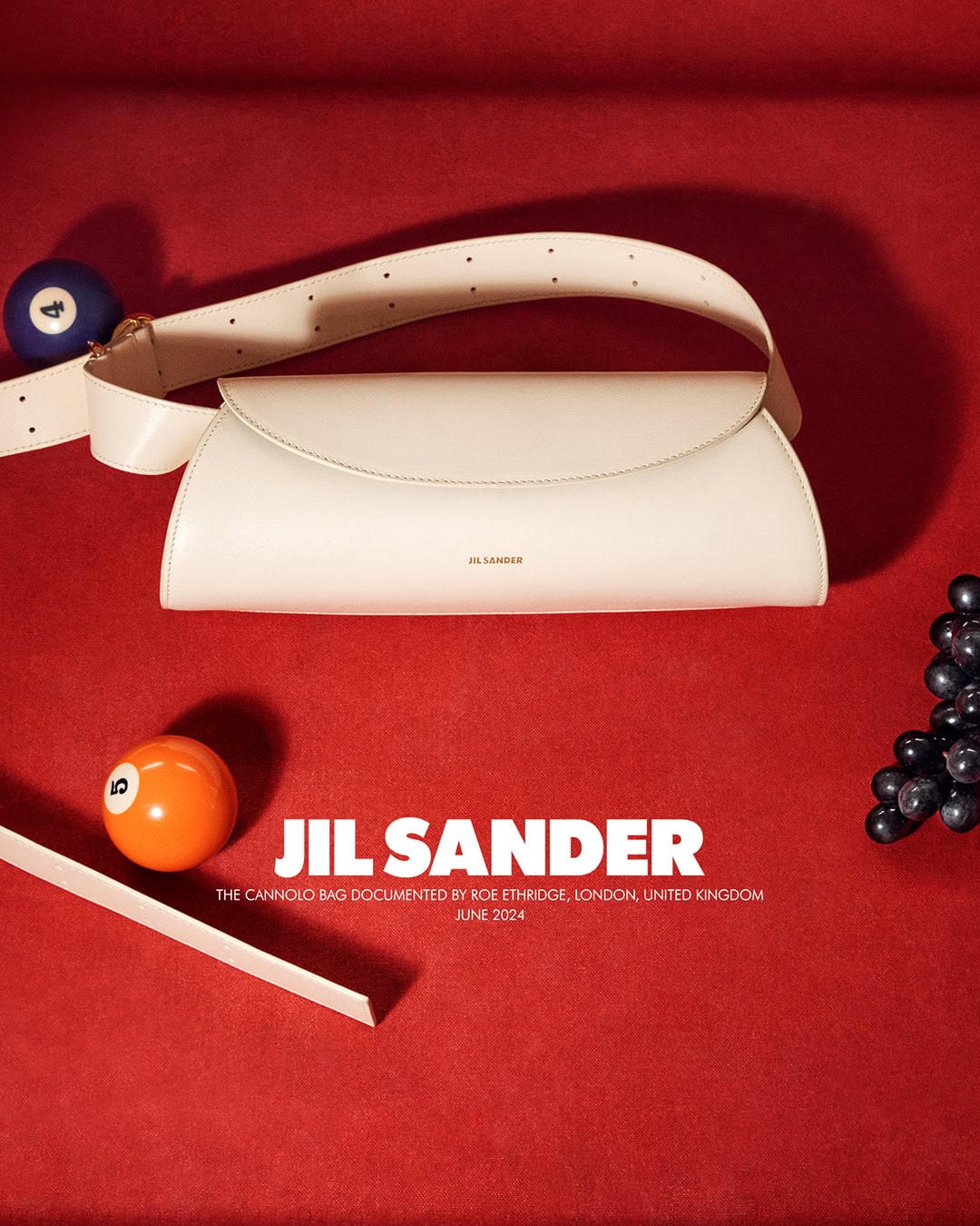 Cream-colored Jil Sander Cannolo bag featured with pool balls and grapes on a red surface, in a product-focused fashion shot.