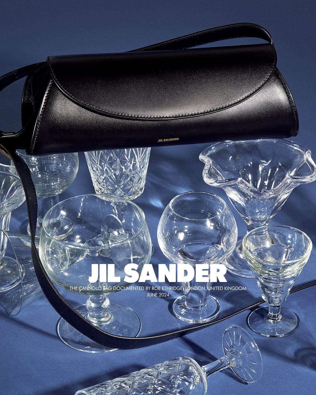 Black Jil Sander handbag displayed on a blue surface surrounded by multiple clear empty crystal glassware with stemmed and footed bases.