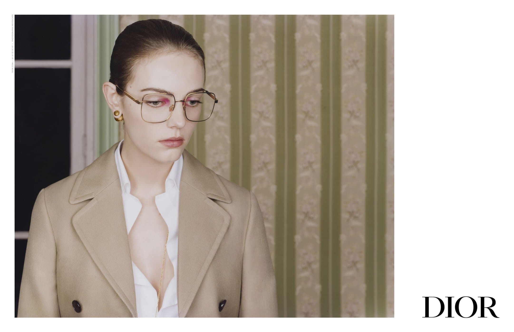 A woman models glasses, wearing a neutral trench coat and collared shirt indoors, against a muted green striped wallpaper backdrop.
