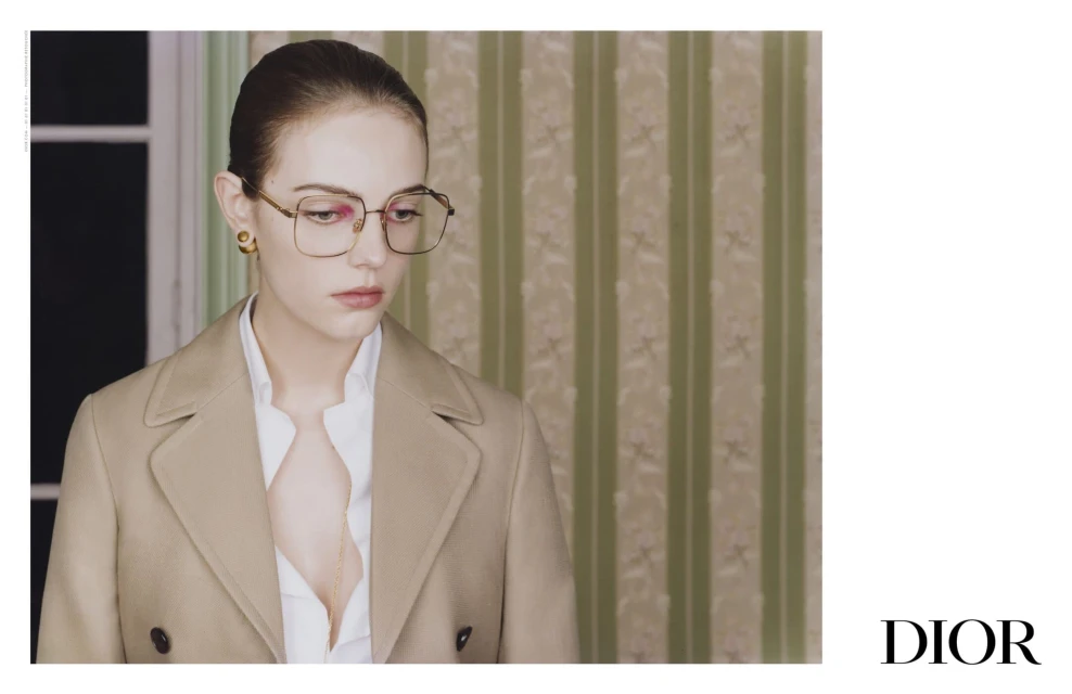 A woman models glasses, wearing a neutral trench coat and collared shirt indoors, against a muted green striped wallpaper backdrop.