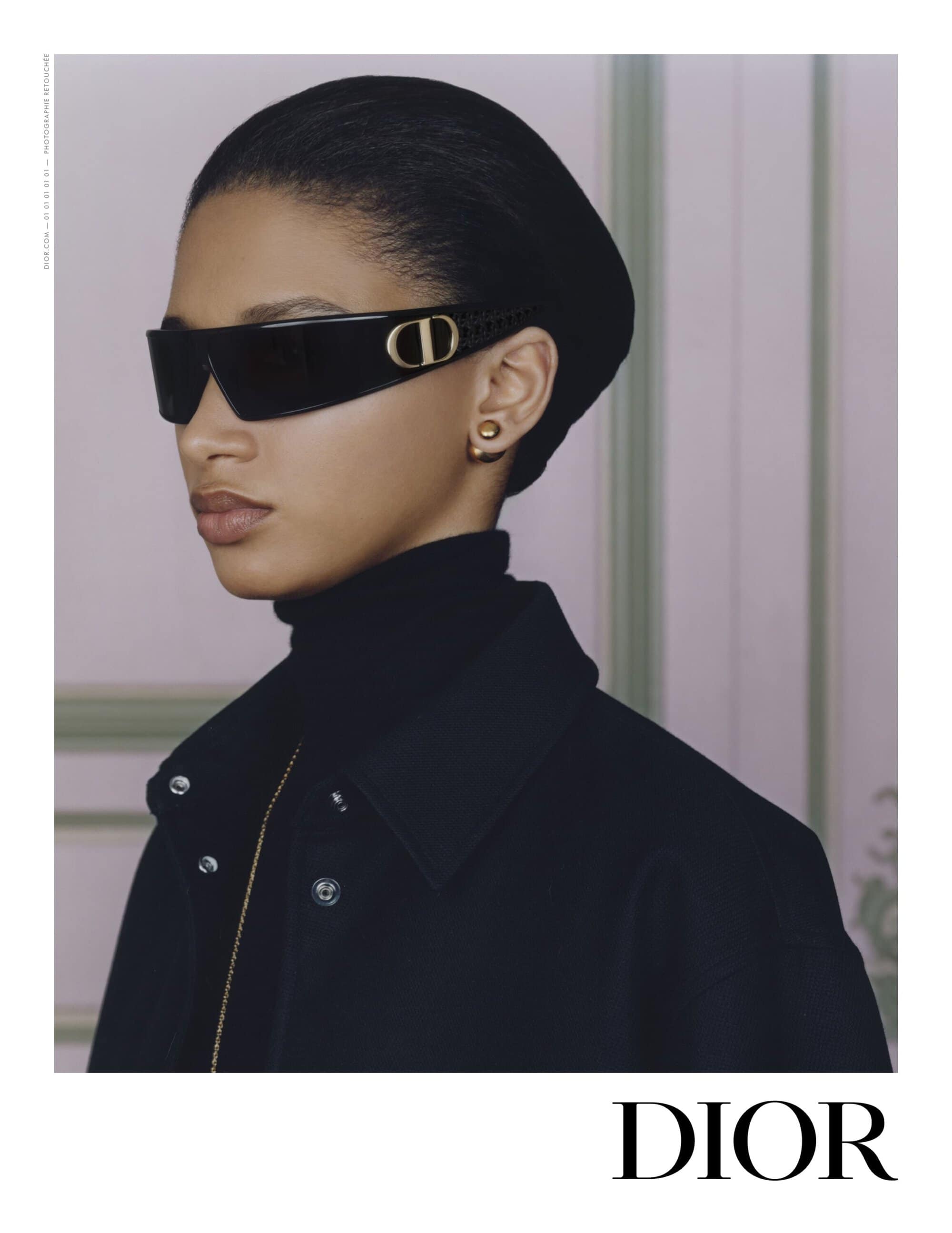 Elegant portrait of a woman wearing black sunglasses with a gold logo, a black hat, and a black coat in a minimalist setting.