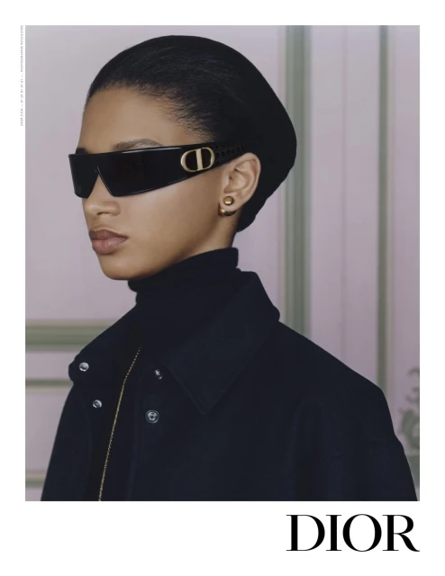 Elegant portrait of a woman wearing black sunglasses with a gold logo, a black hat, and a black coat in a minimalist setting.