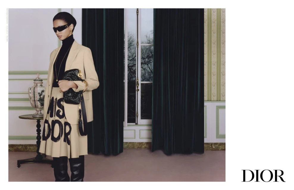 Fashion editorial shot of a woman in a blazer, skirt, and sunglasses holding a handbag, posing in an indoor setting near a window.
