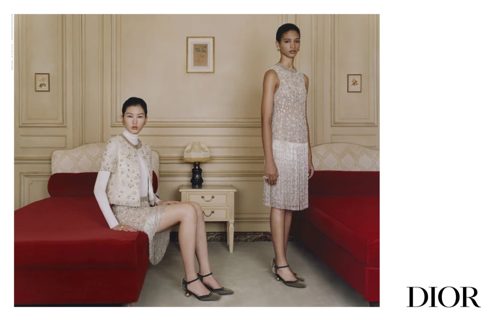 Two women in elegant, beaded dresses pose in an indoor setting with red couches, showcasing a blend of fashion and interior design.