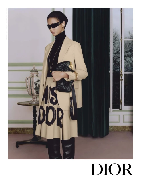 Fashion editorial shot of a slender woman wearing a tan blazer, black turtleneck, tan skirt with black text, sunglasses, and black boots.