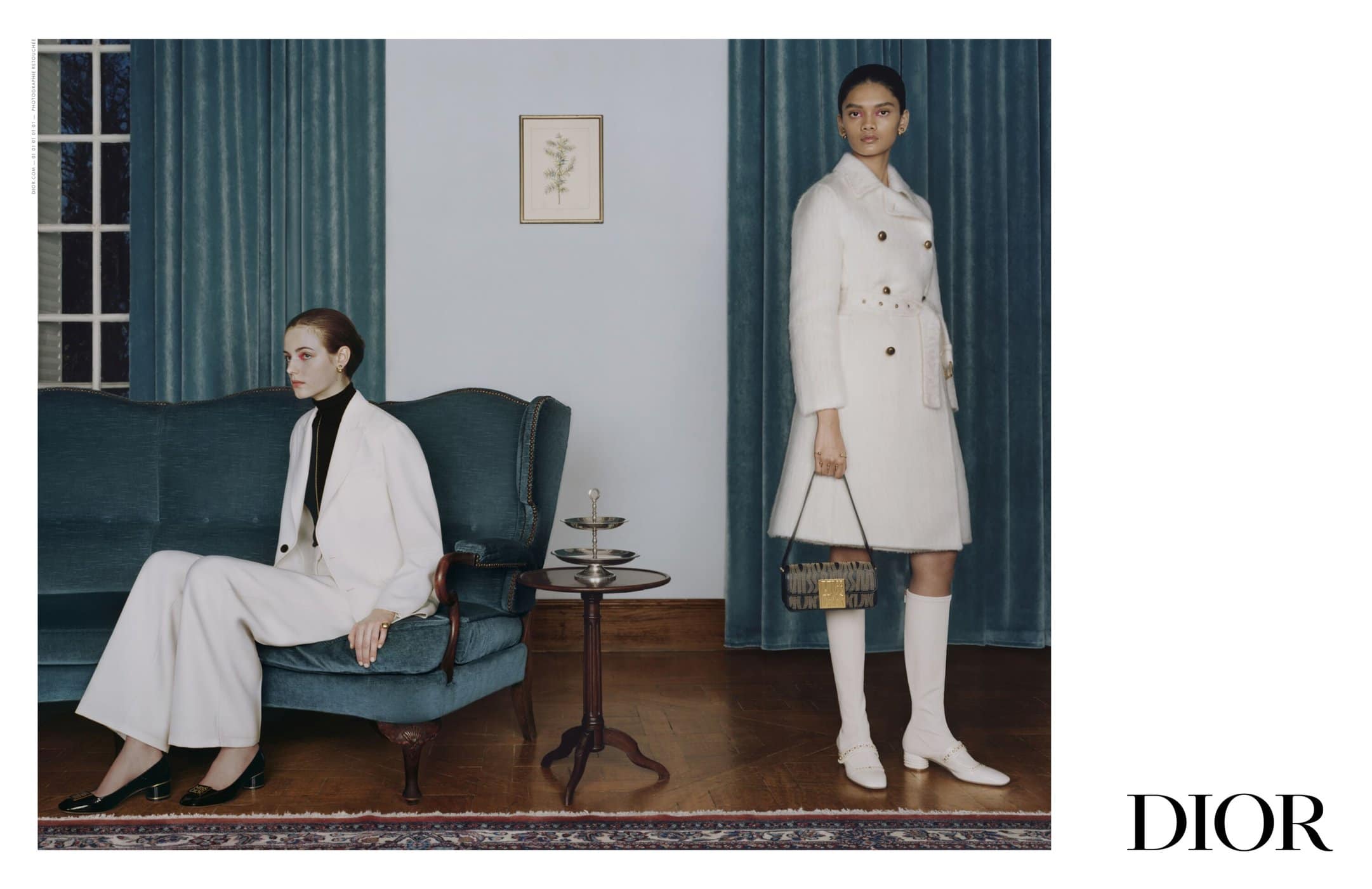 Two women in white coats and suits pose in an indoor setting with a teal sofa and wooden floor, accented by a dark-colored handbag and 'DIOR' logo.