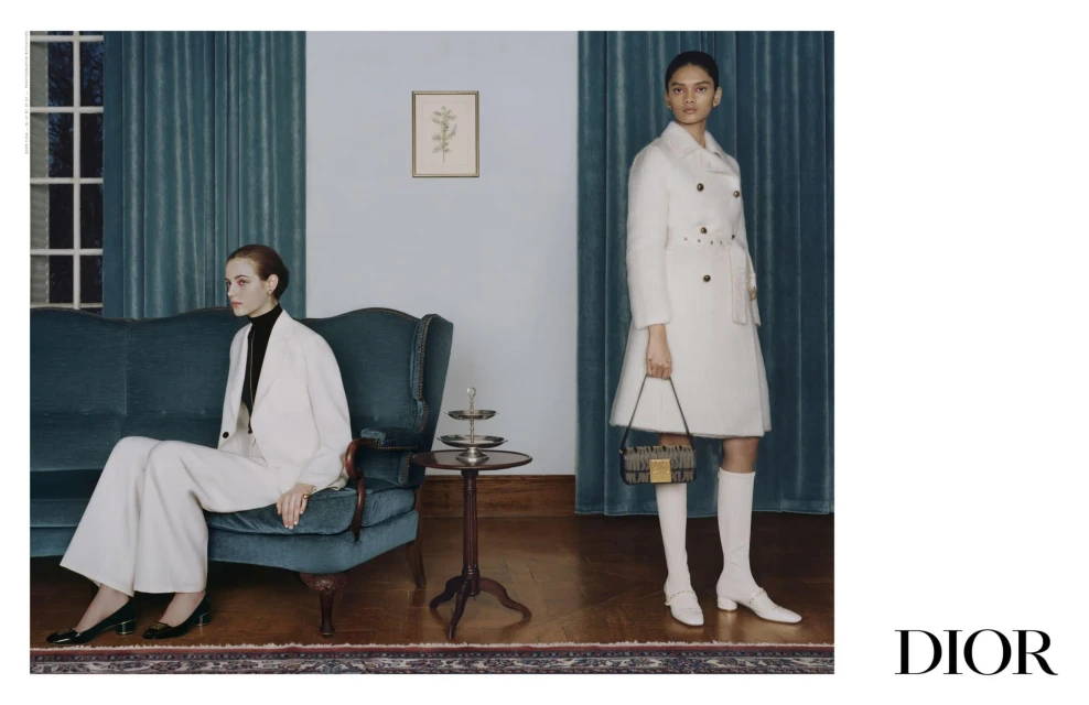 Two women in white coats and suits pose in an indoor setting with a teal sofa and wooden floor, accented by a dark-colored handbag and 'DIOR' logo.