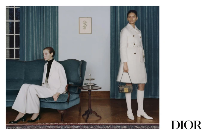 Two women in white coats and suits pose in an indoor setting with a teal sofa and wooden floor, accented by a dark-colored handbag and 'DIOR' logo.