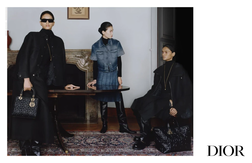 Three women model black and denim outfits and purses indoors, posing near a wooden table and fireplace, with a 'DIOR' logo visible.