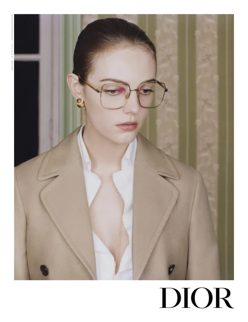 Portrait of a young woman with auburn hair and glasses wearing a tan jacket and white shirt, in front of a striped wallpaper.