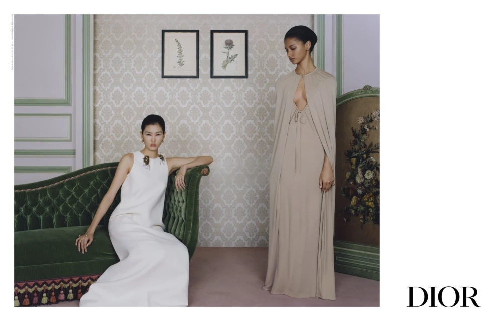 Two women in elegant gowns pose in a vintage-styled room with floral wallpaper, a green velvet couch, and framed botanical art.