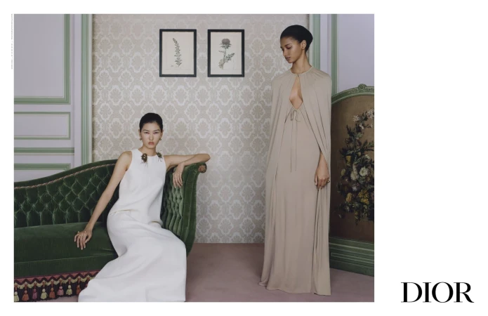 Two women in elegant gowns pose in a vintage-styled room with floral wallpaper, a green velvet couch, and framed botanical art.