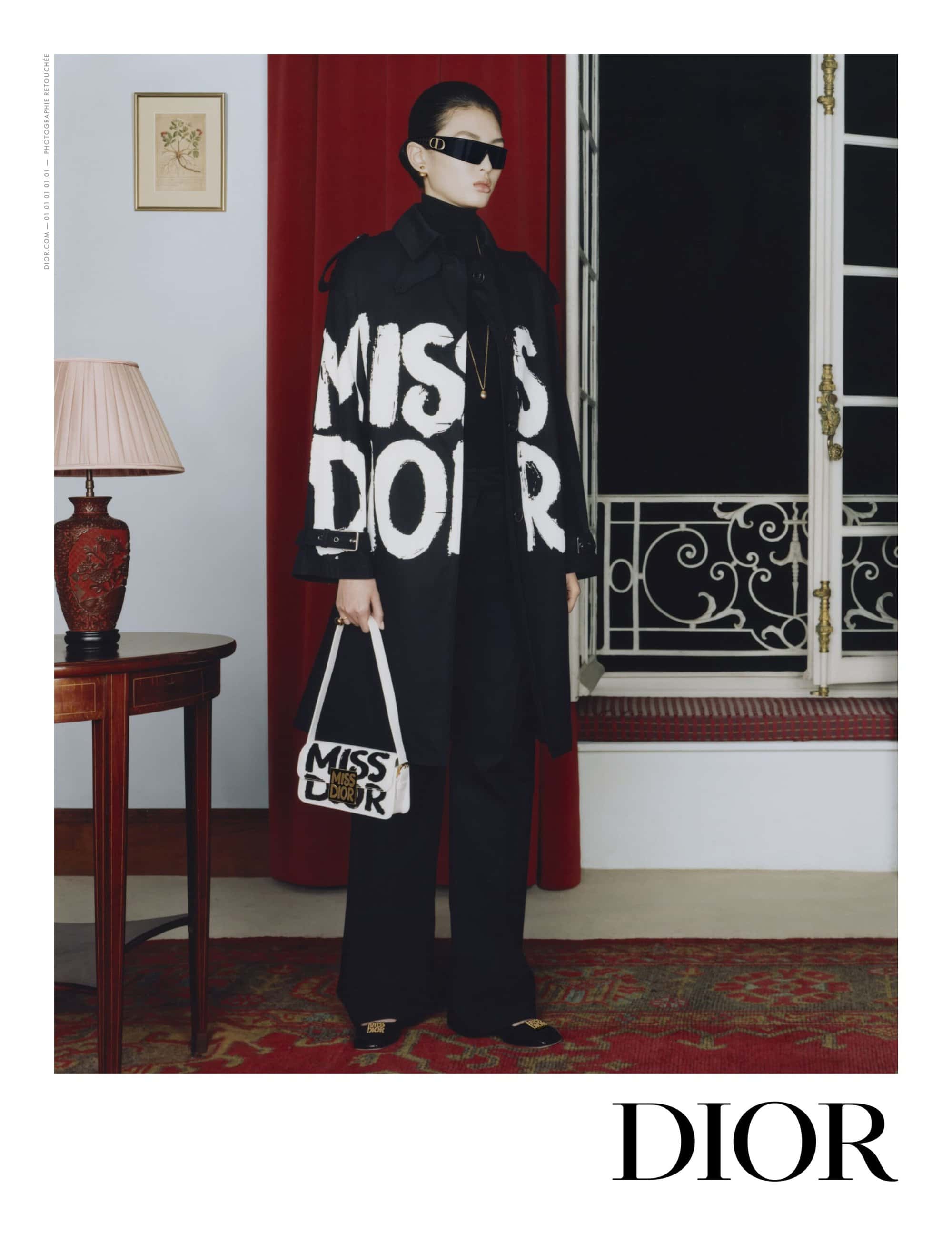 Model wearing a black Miss Dior trench coat and sunglasses, holding a white logo handbag, standing in a classic room with a red curtain.