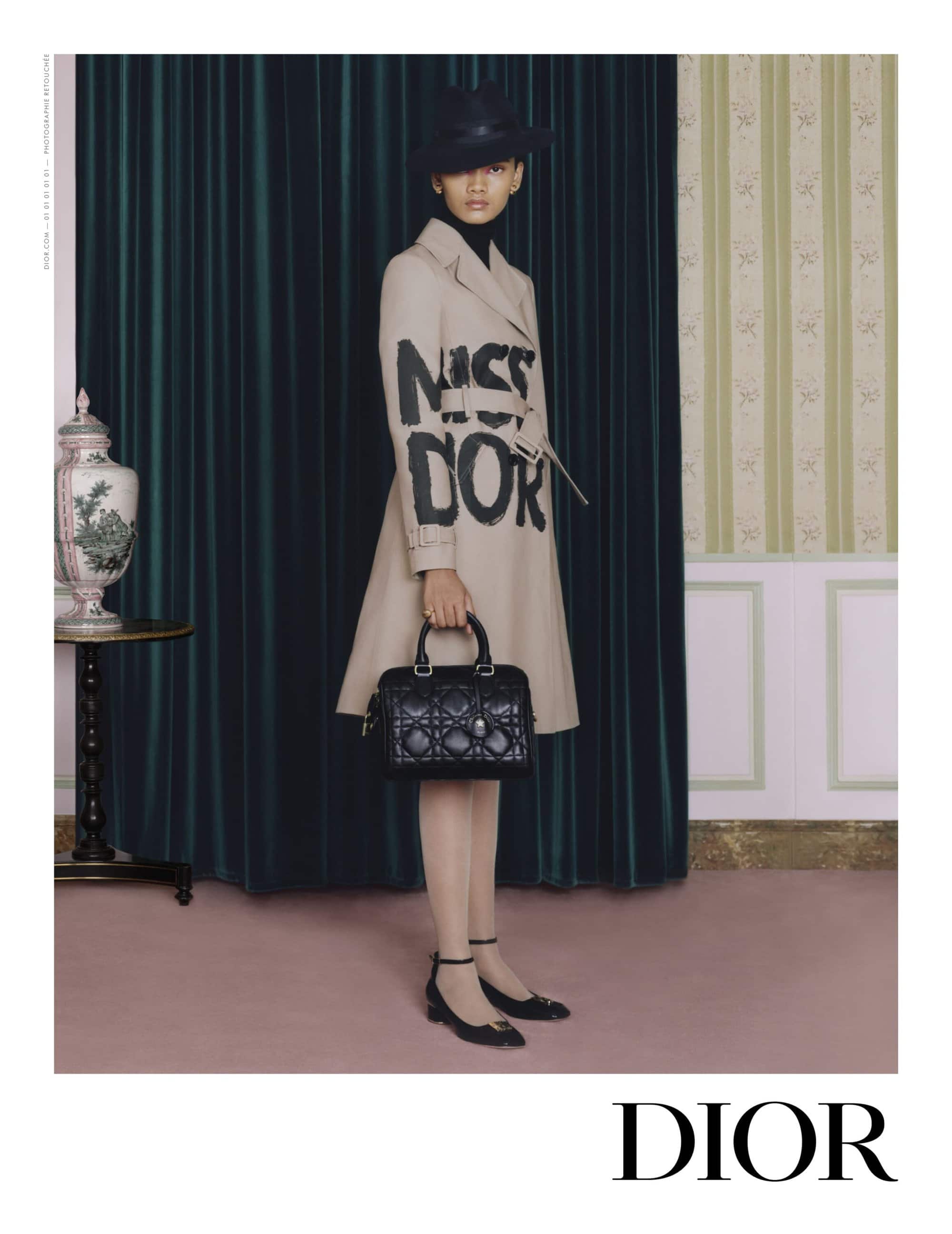 Woman in a beige trench coat with 'Miss Dior' lettering, holding a black quilted handbag, posing against a classic curtain and wall.