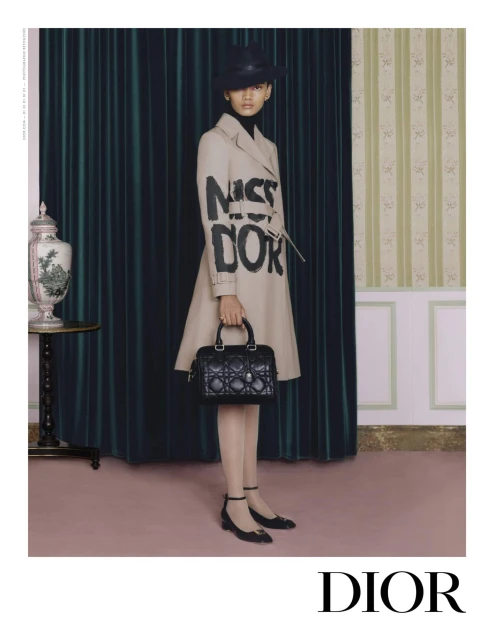 Woman in a beige trench coat with 'Miss Dior' lettering, holding a black quilted handbag, posing against a classic curtain and wall.