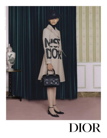 Woman in a beige trench coat with 'Miss Dior' lettering, holding a black quilted handbag, posing against a classic curtain and wall.