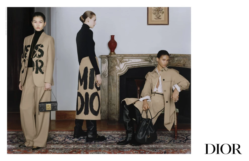 Three women model neutral-toned Dior outfits inside a room with a fireplace and ornate rug for a fashion editorial shot.