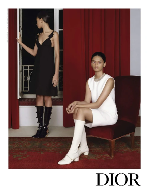 Two women model elegant dresses and knee-high boots in a refined indoor setting with rich textures, bathed in soft, ambient light.