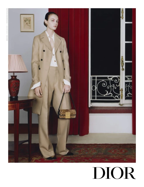 A woman in a beige pantsuit and trench coat poses indoors with a leopard print purse, next to a vintage desk, red curtains and window.