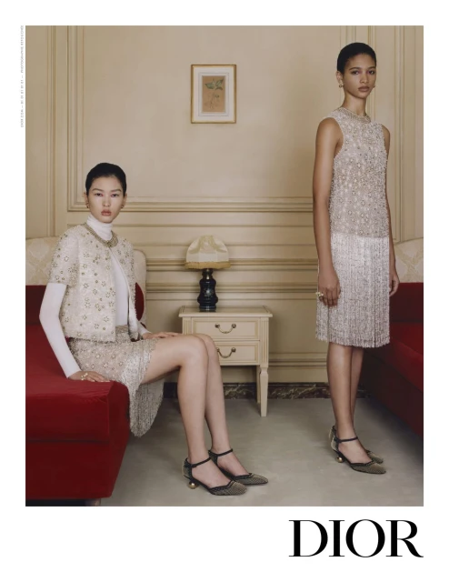Two elegant Asian women showcase couture fashion indoors. They are wearing ornate embellished skirt suits by Dior, posing in front of a wall.