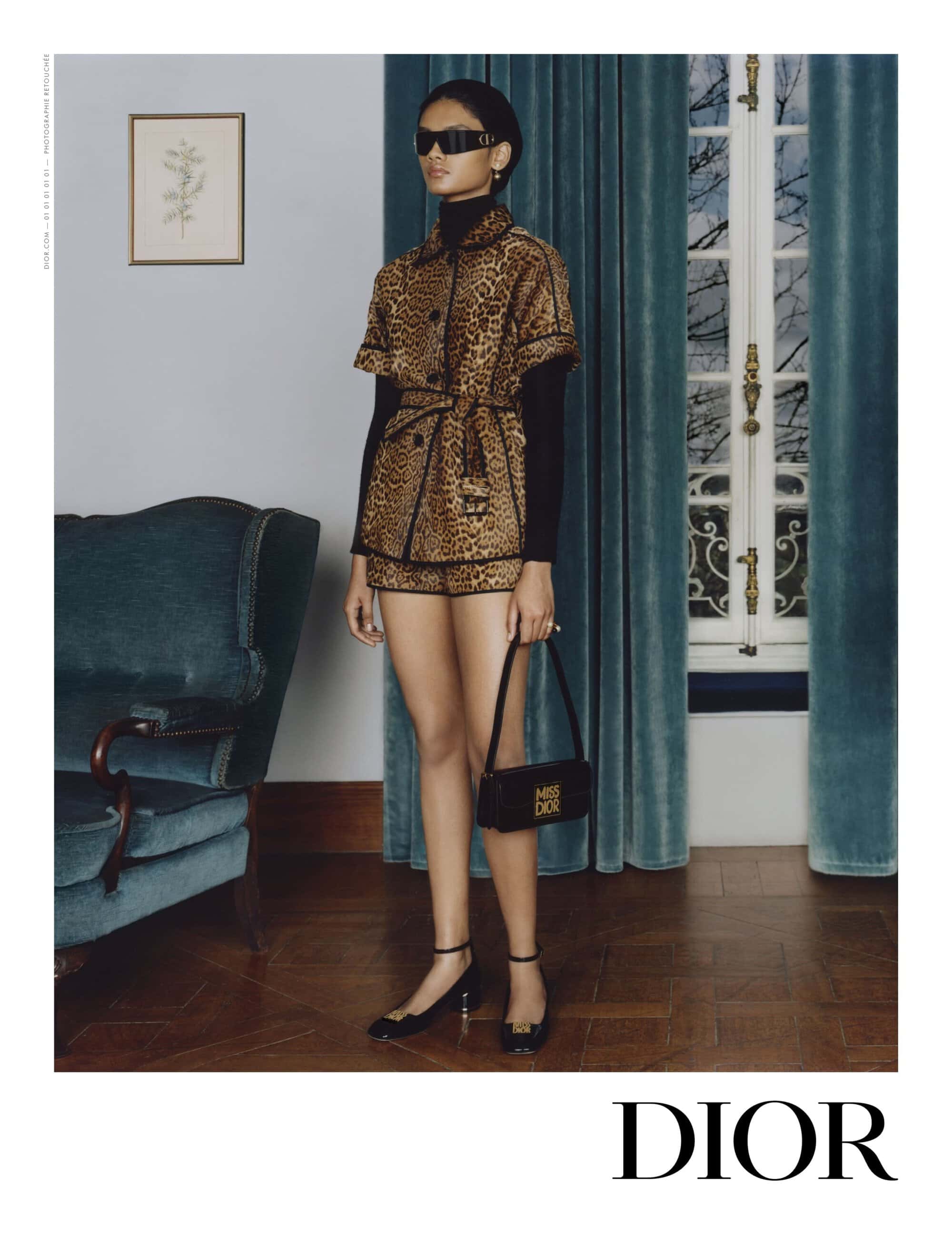 Woman in a leopard print shorts suit with black top, sunglasses, and handbag, posing in a room with a vintage chair.