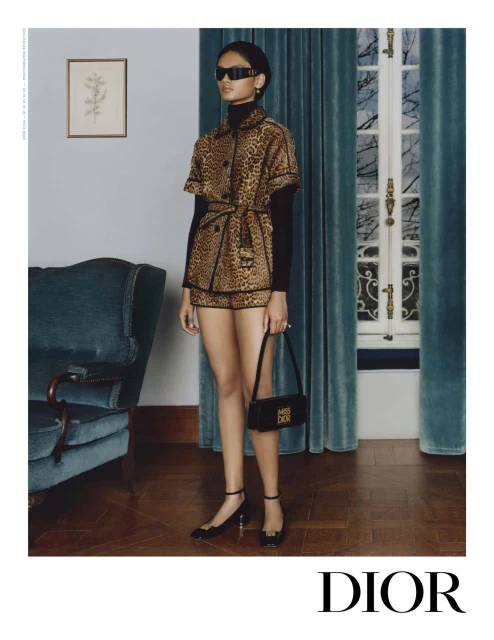 Woman in a leopard print shorts suit with black top, sunglasses, and handbag, posing in a room with a vintage chair.