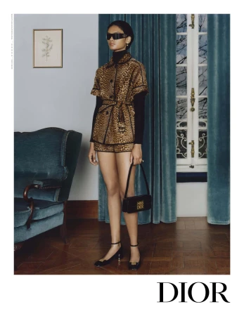 Woman in a leopard print shorts suit with black top, sunglasses, and handbag, posing in a room with a vintage chair.