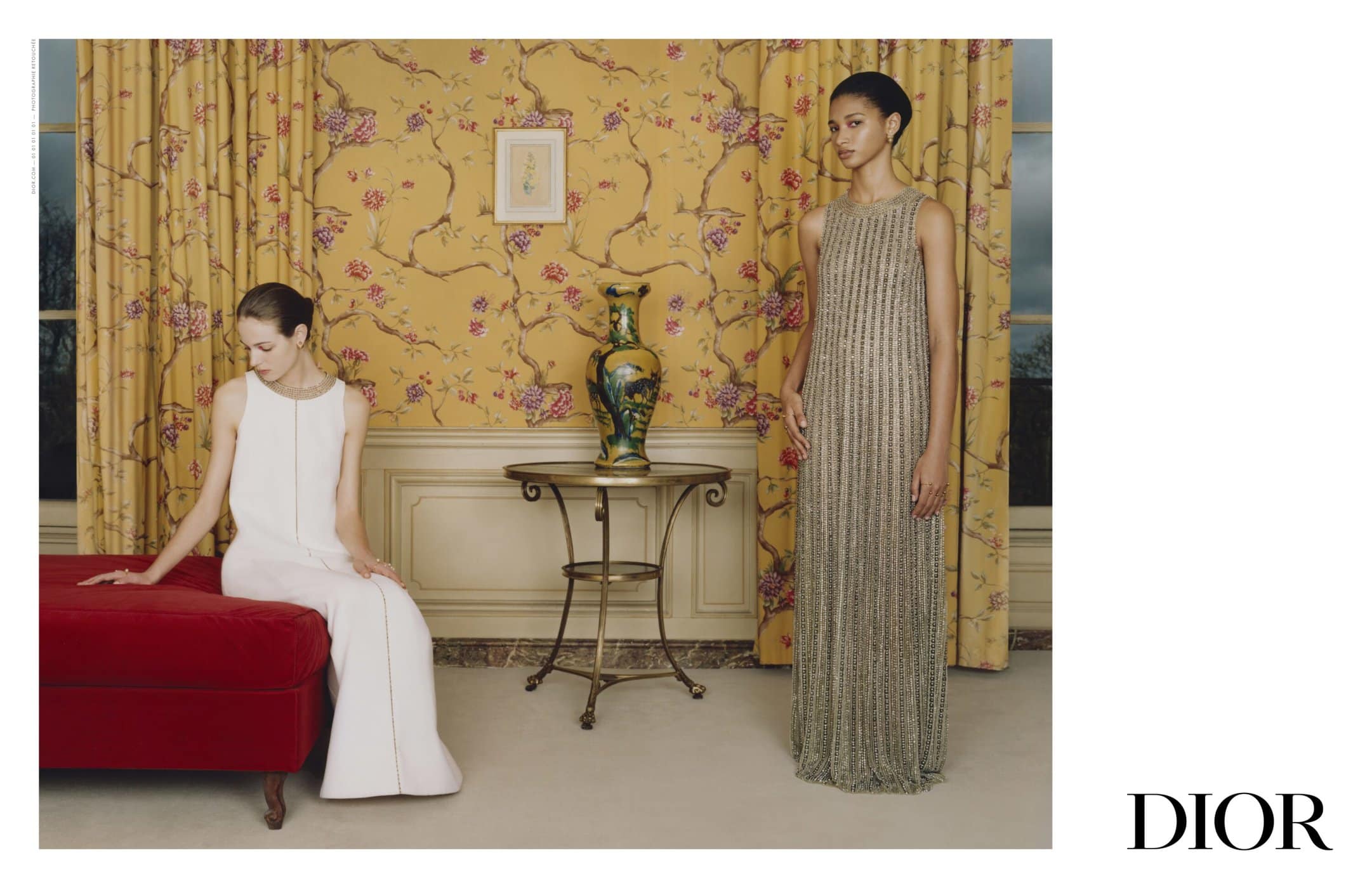 Two fashion models in elegant gowns pose indoors against a yellow floral wallpaper backdrop, beside vintage furniture and the Dior logo.